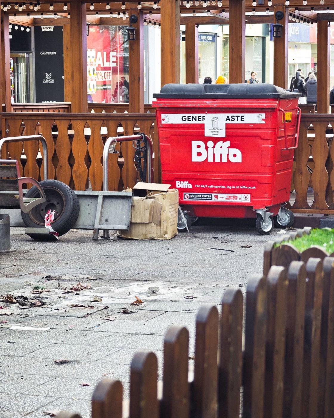 A red Biffa general waste bin behind the market fence, litter on the ground
